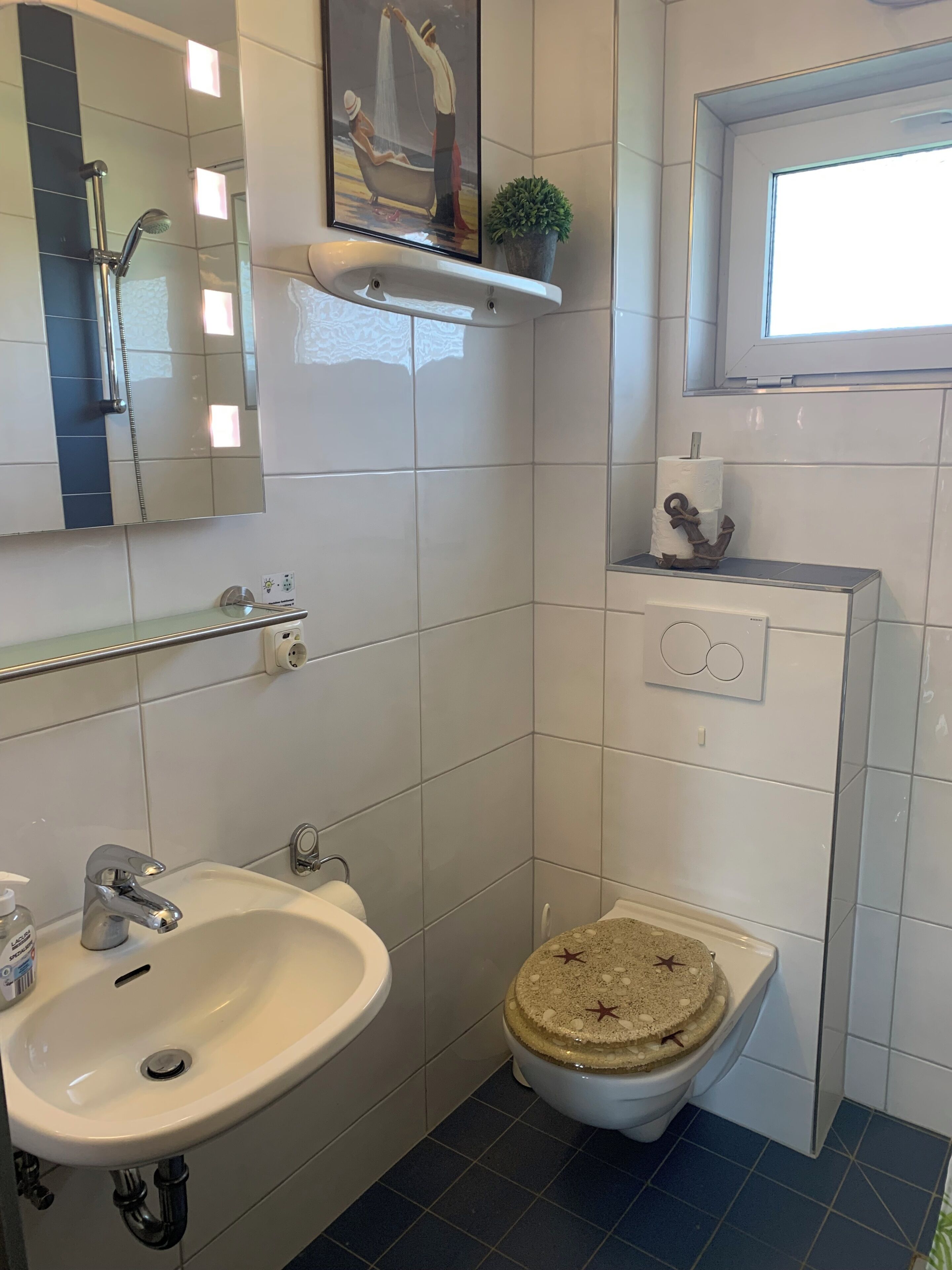 Un baño con lavamanos blanco, inodoro con tapa decorativa, zona de ducha y ventana.