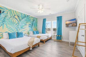Ett hotellrum med två sängar, en takfläkt, ett stort fönster med gardiner, en tavla på väggen och en trästege.