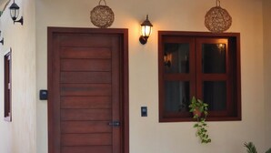 Eine Holztür mit einem Metallgriff, ein Fenster mit hängenden Pflanzen darunter und zwei Wandleuchten.