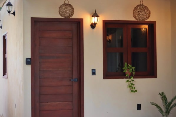 A wooden door with a metal handle, a window with a plant hanging below it, and two wall-mounted light fixtures.