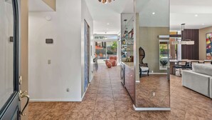 Um interior moderno com um corredor que dá para uma sala de jantar, uma cozinha com armários de vidro e uma sala de estar com sofá e poltronas.