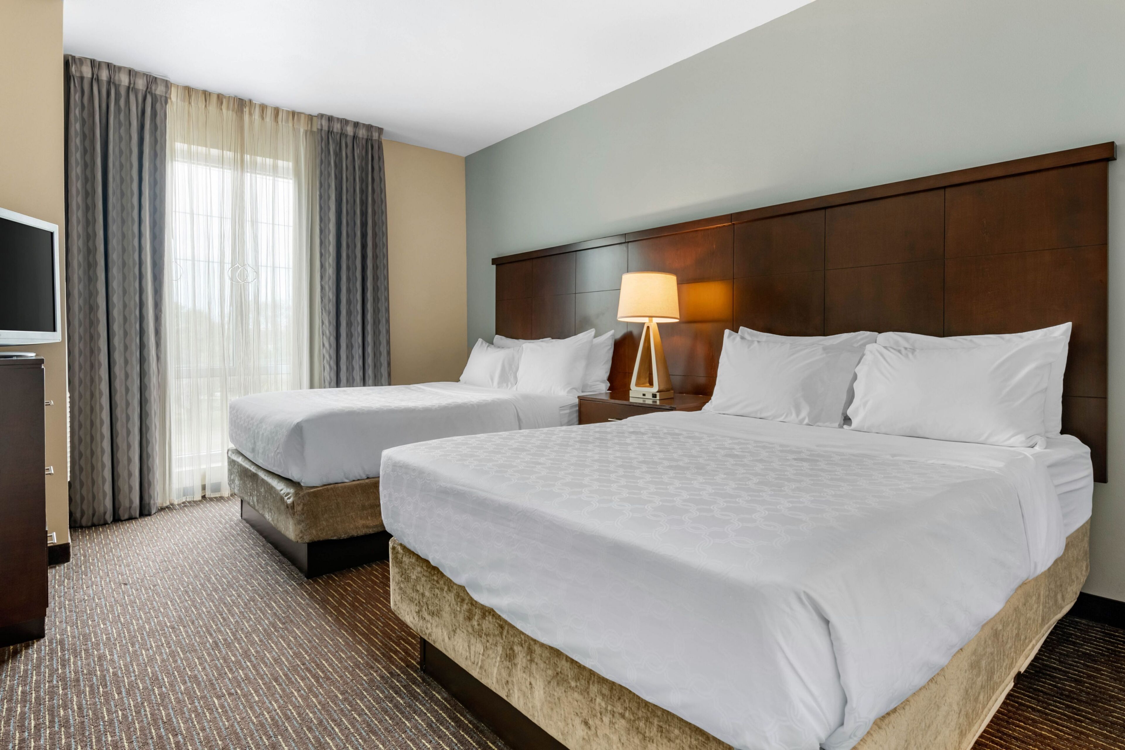 A hotel room with two beds, a television, a lamp, and a large window with curtains.
