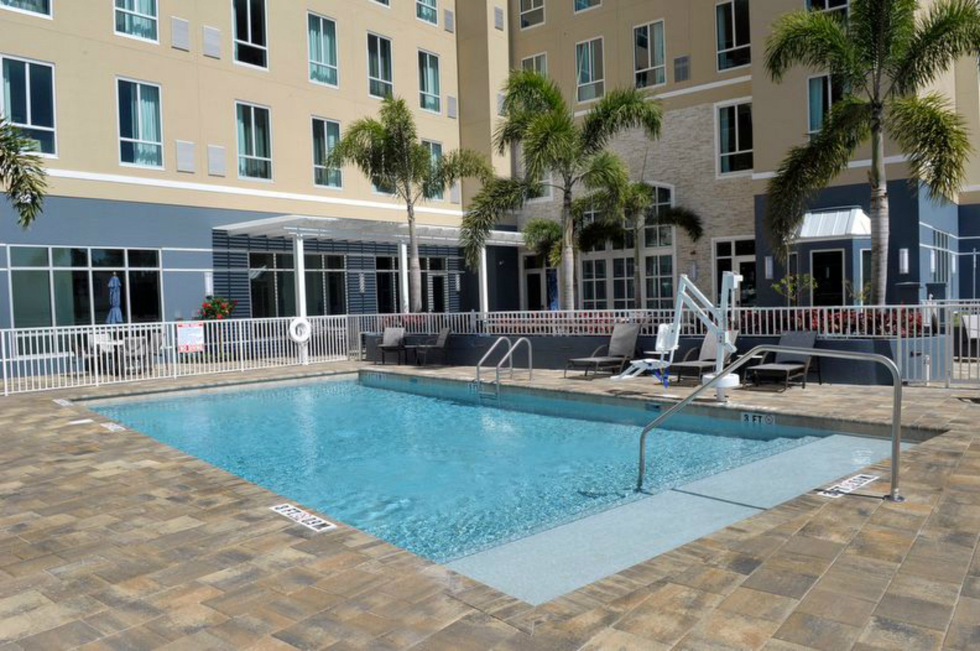 A swimming pool with a tiled deck, surrounded by a white fence, and palm trees.