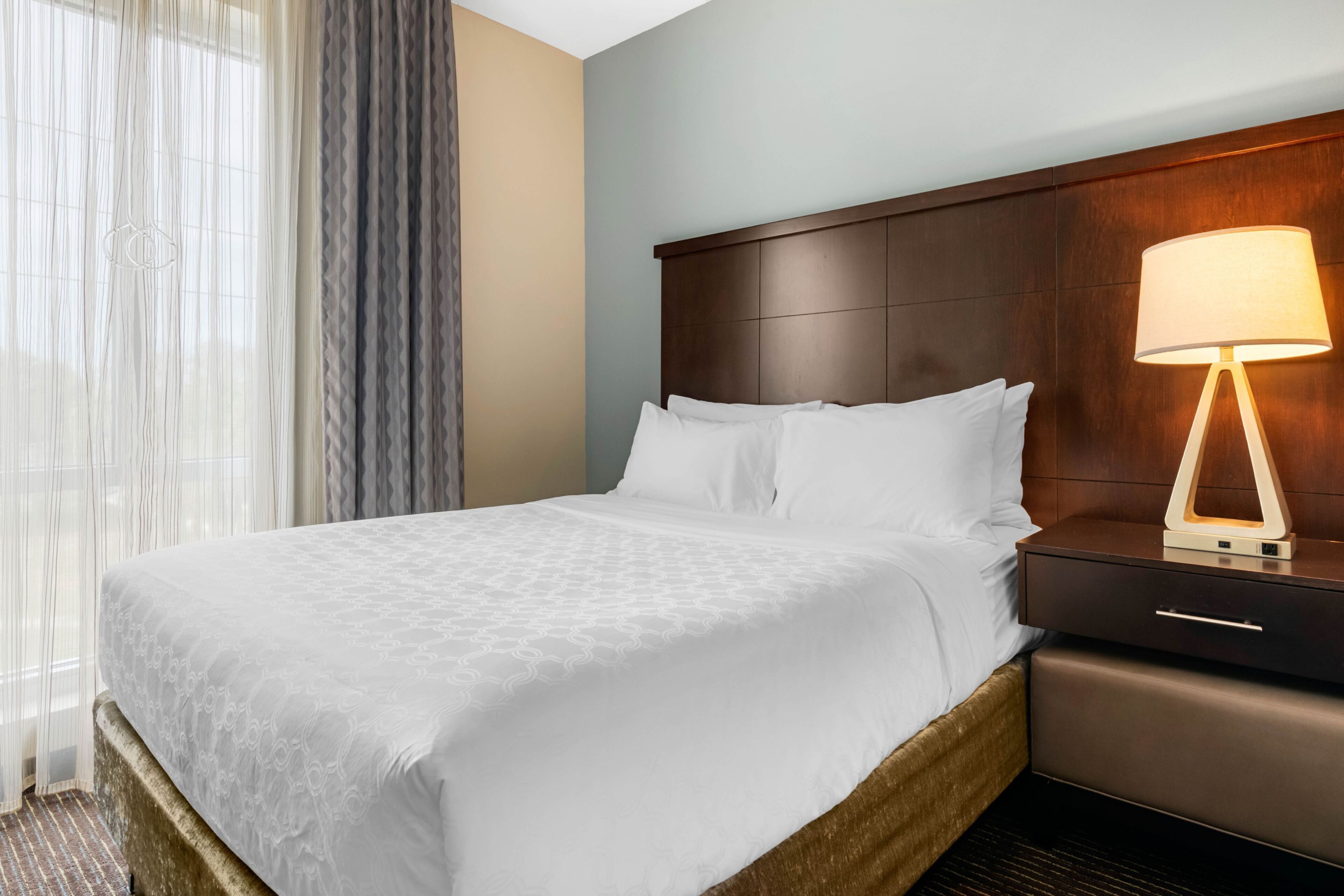 A hotel room with a large bed, a bedside table with a lamp, and a window with sheer curtains.