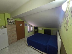 A small, single-room apartment with a sloped ceiling, a bed with a blue cover, a white refrigerator, a wooden cabinet, and a door. - Vrbo Property (Riposto)