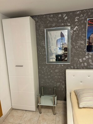 Een slaapkamer met een witte kast, een glazen tafel, een bed met een gestreept kussen en een lijst met een afbeelding van een gebouw.
