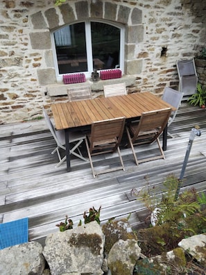 A wooden deck with a stone wall, a table with chairs, and a window with a view. - Charming stone house with a garden – Finistère, near the sea (Quéménéven)