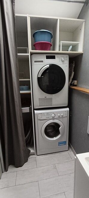 A compact laundry area with a stacked washer and dryer, a shelf with a blue container, and a curtain partially covering the space. - Charming stone house with a garden – Finistère, near the sea (Quéménéven)