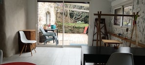 A room with a sliding glass door leading to an outdoor area with a stone wall and greenery. Inside, there is a table with chairs, a wooden cabinet, and a ladder against the wall. - Charming stone house with a garden – Finistère, near the sea (Quéménéven)