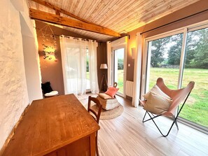 A cozy room with a wooden table, a cushioned chair, and a view of a grassy yard through large windows.