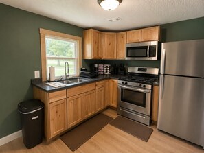 A kitchen with wooden cabinets, stainless steel appliances, a window, and a trash bin. - Bluebird Cabin: The Scenic Hills Cottages (Logan)