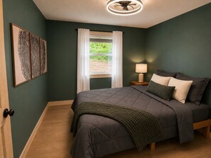 A bedroom with a bed, a bedside table, a lamp, and a window with curtains. - Bluebird Cabin: The Scenic Hills Cottages (Logan)