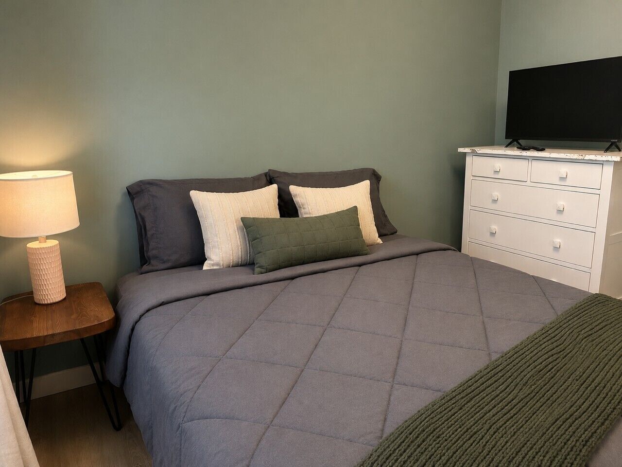 A bedroom with a bed, a nightstand with a lamp, a dresser, and a television.
