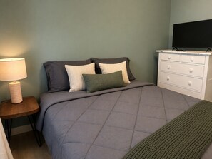 A bedroom with a bed, a nightstand with a lamp, a dresser, and a television. - Bluebird Cabin: The Scenic Hills Cottages (Logan)
