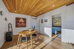 Une pièce avec un plafond en bois, une table à manger avec des chaises, un lit recouvert d’une couverture bleue et une fenêtre donnant sur des arbres.