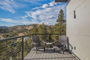 Un balcón con dos sillas Adirondack y una pequeña mesa redonda, con vistas a un paisaje montañoso con árboles y cielo despejado.
