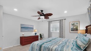 Une chambre à coucher comprenant un lit, une table de chevet, un ventilateur de plafond, une fenêtre avec des stores et un tableau accroché au mur.