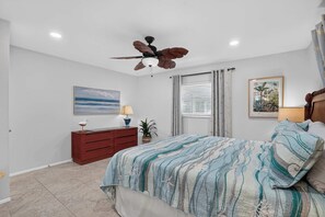 Ein Schlafzimmer mit Bett, Nachttisch, Deckenventilator, Fenster mit Jalousien und einem Bild an der Wand.