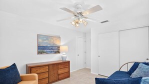 Une pièce avec un ventilateur de plafond, un tableau représentant un paysage marin, une armoire en bois, une lampe et un lit avec une literie bleue.