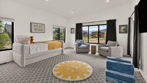 Ein modernes Schlafzimmer mit einem Bett, zwei Sesseln, einem kleinen Tisch und einem großen Fenster mit Blick ins Freie.
