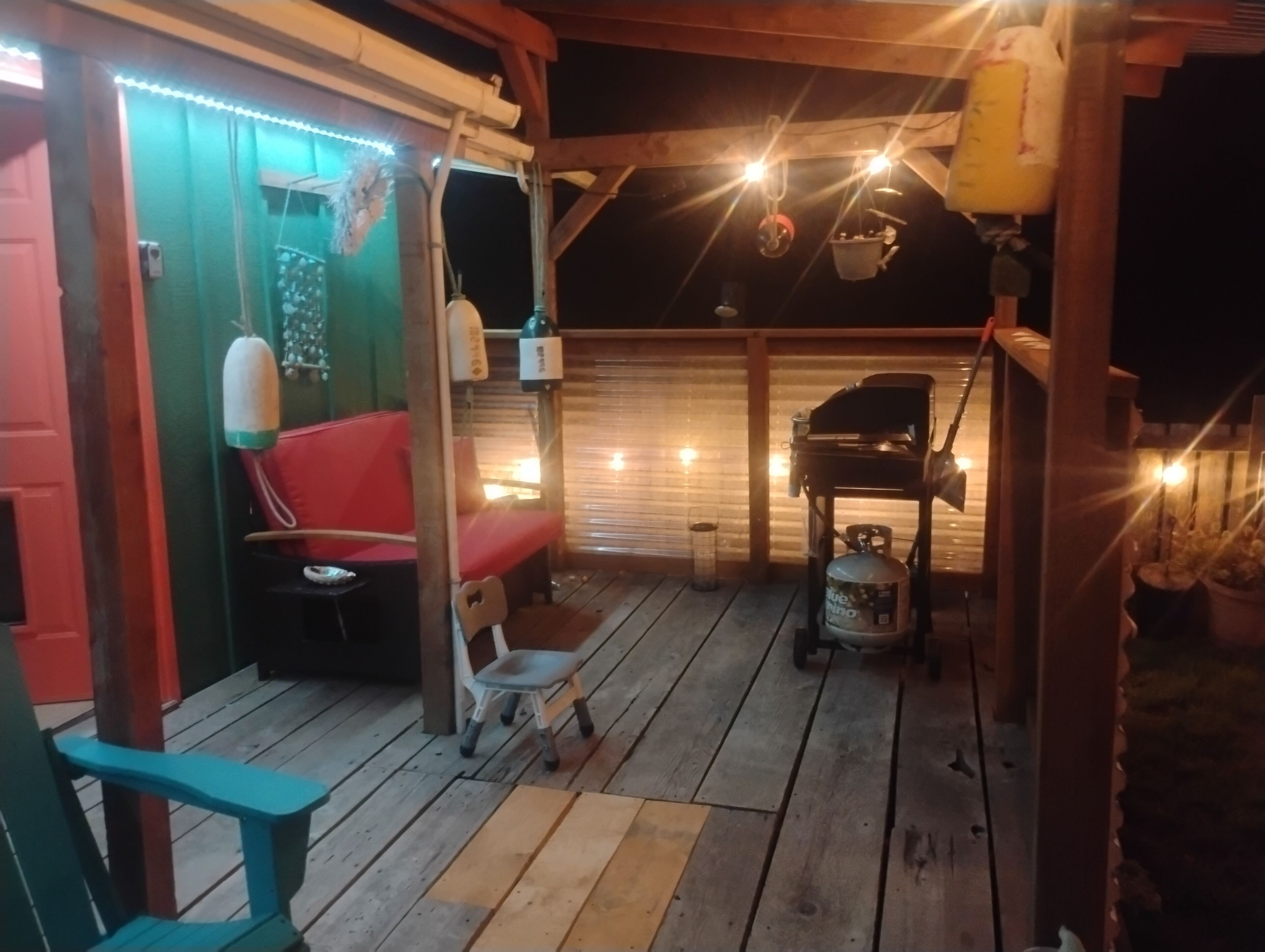 A covered outdoor area with a wooden deck, a red bench, a white chair, a gas cylinder, and a barbecue grill.