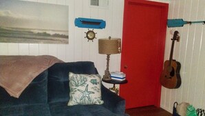 A room with a red door, a blue couch with a starfish pillow, a small table with a lamp, and a guitar leaning against the wall. - Peaceful 1-bedroom cabin in adorable Moclips perfect for relaxing (Moclips)