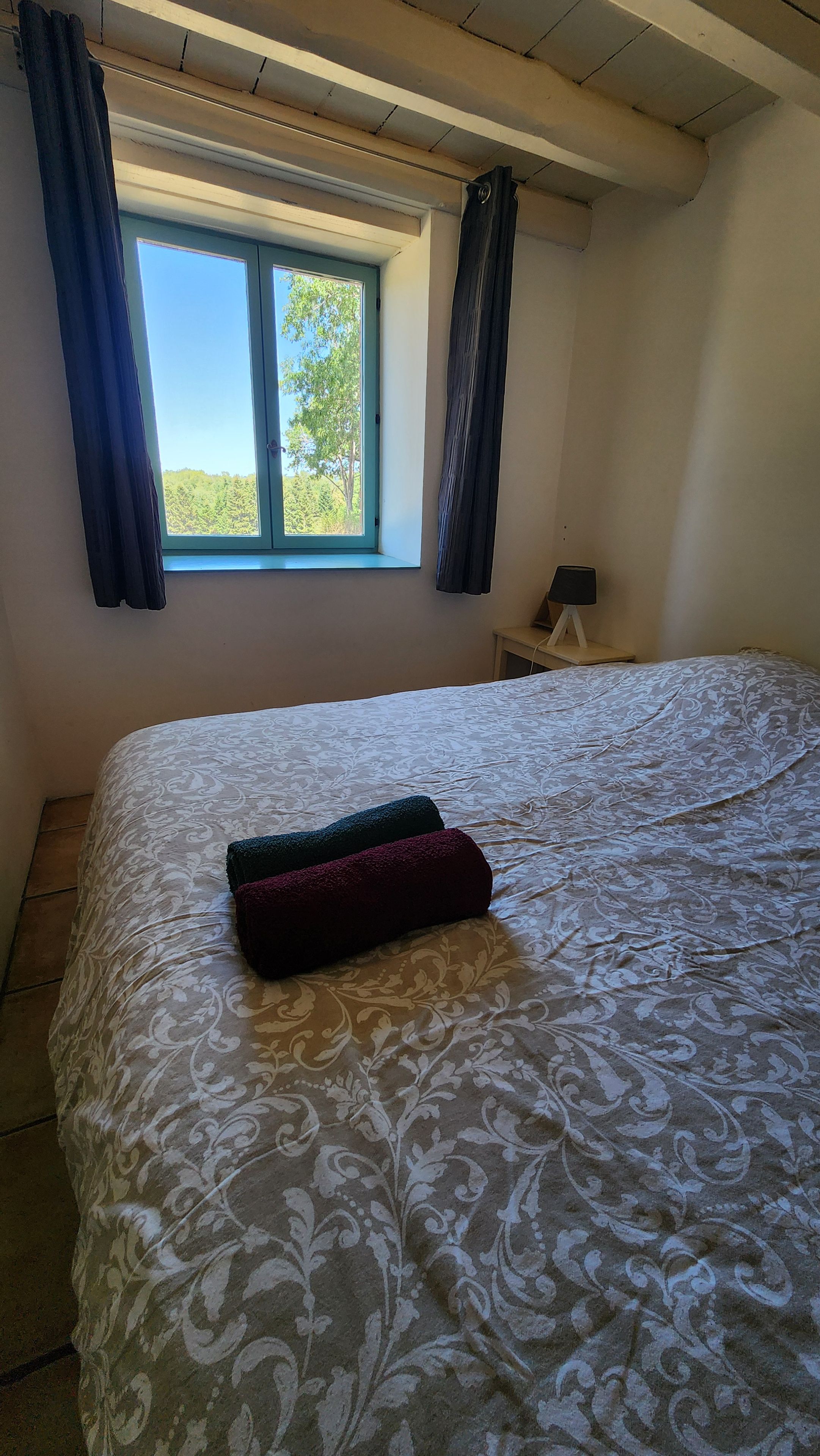 A bedroom with a large bed, a window with curtains, and a small bedside table with a lamp.