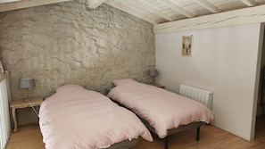 A room with two beds, a rustic stone wall, a radiator, and a small table with a lamp.