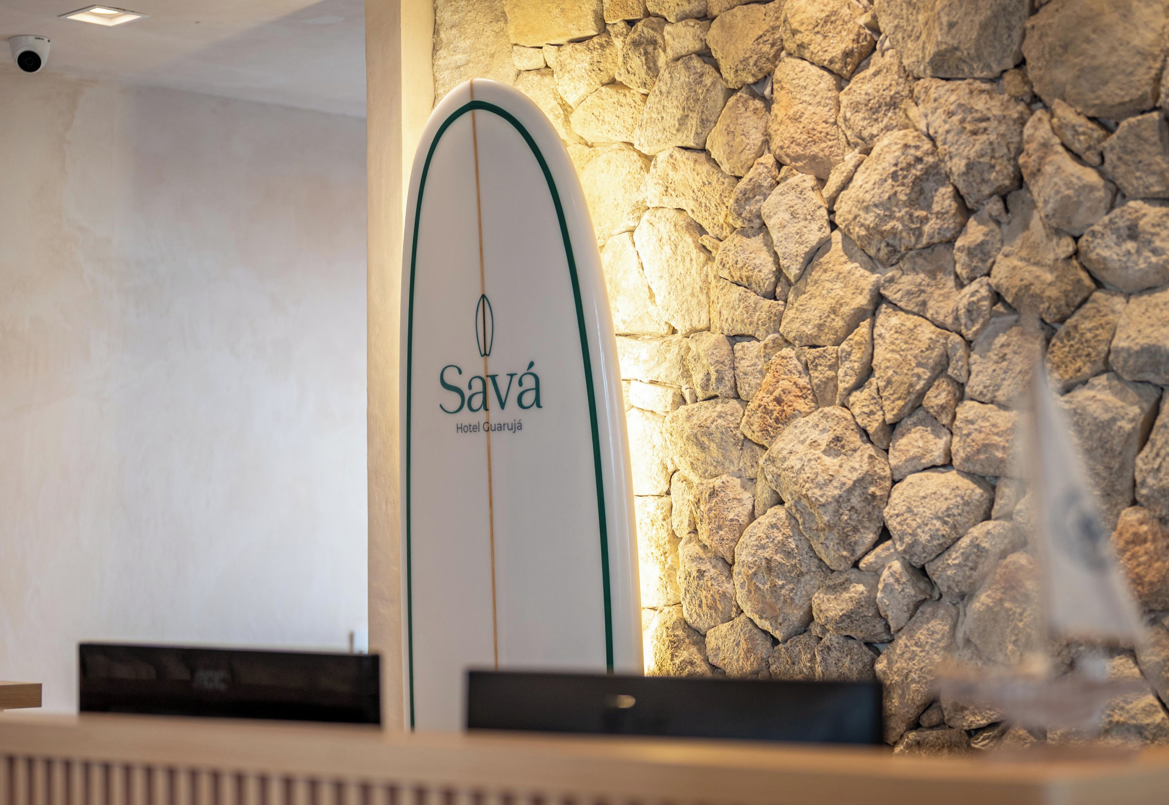 Una recepción con un letrero con forma de tabla de surf que dice 'Sava Hotel Guaruja' sobre un fondo de pared de piedra.