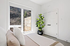 Ein Zimmer mit großem Fenster, weißer Tür, einem Topf mit Pflanze und einem weißen, flauschigen Kissen auf einem Bett.