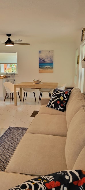 Ein Wohnzimmer mit einem beigefarbenen Sofa, einem Esstisch mit Stühlen, einem Deckenventilator und einem Wandbild im Strandhausstil.