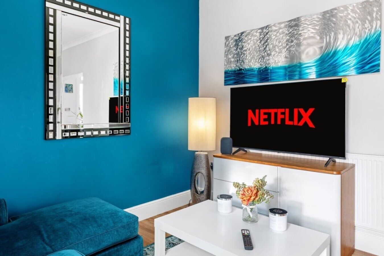 A living room with a blue wall, a large mirror, a flat-screen TV displaying 'NETFLIX', a white coffee table with a vase and candles, and a blue sofa.