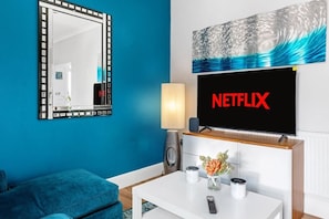 Ein Wohnzimmer mit blauer Wand, einem großen Spiegel, einem Flachbildfernseher, der 'NETFLIX' zeigt, einem weißen Couchtisch mit einer Vase und Kerzen sowie einem blauen Sofa.
