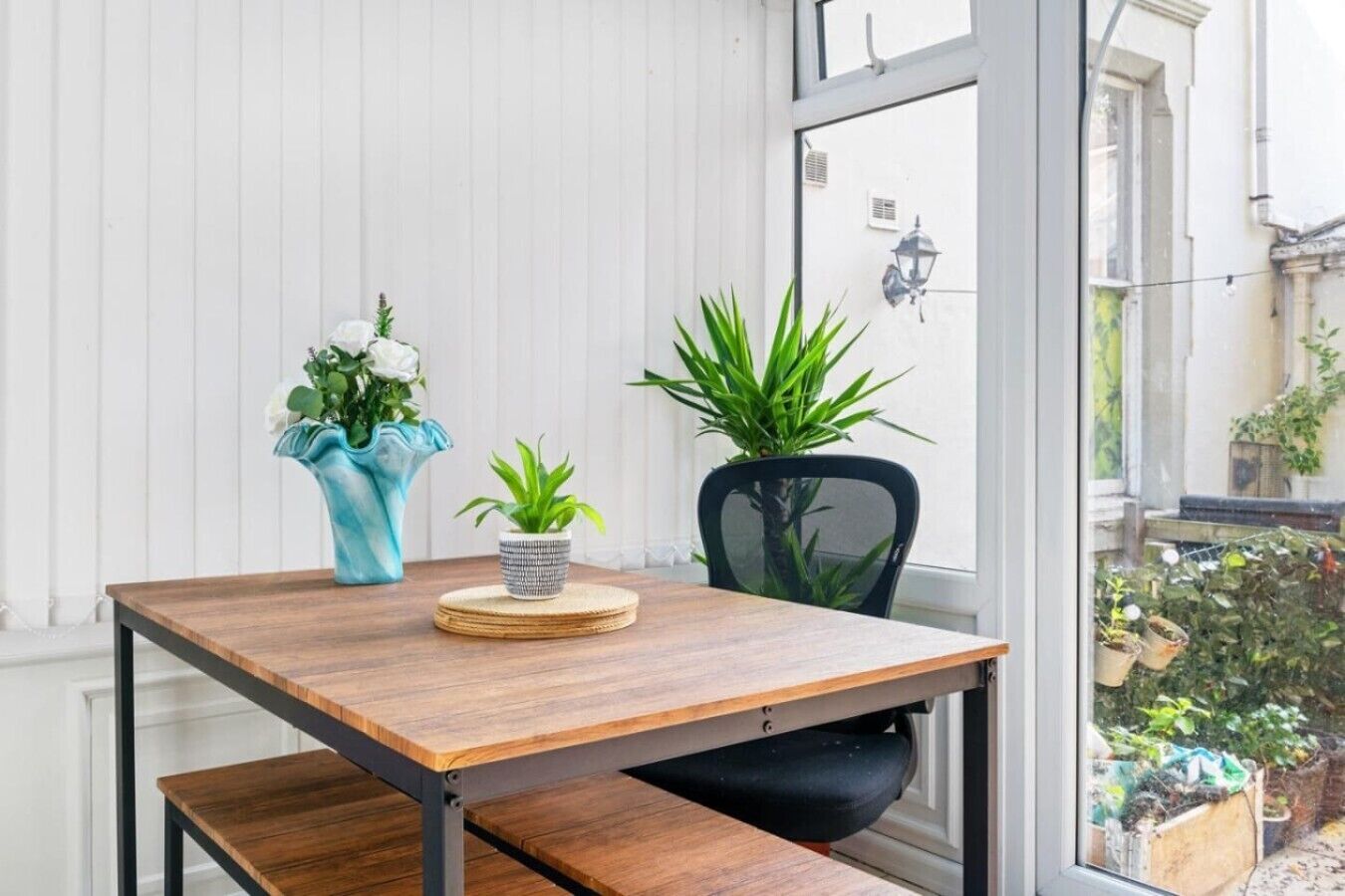 A wooden table with a bench, two potted plants, and a vase with flowers.