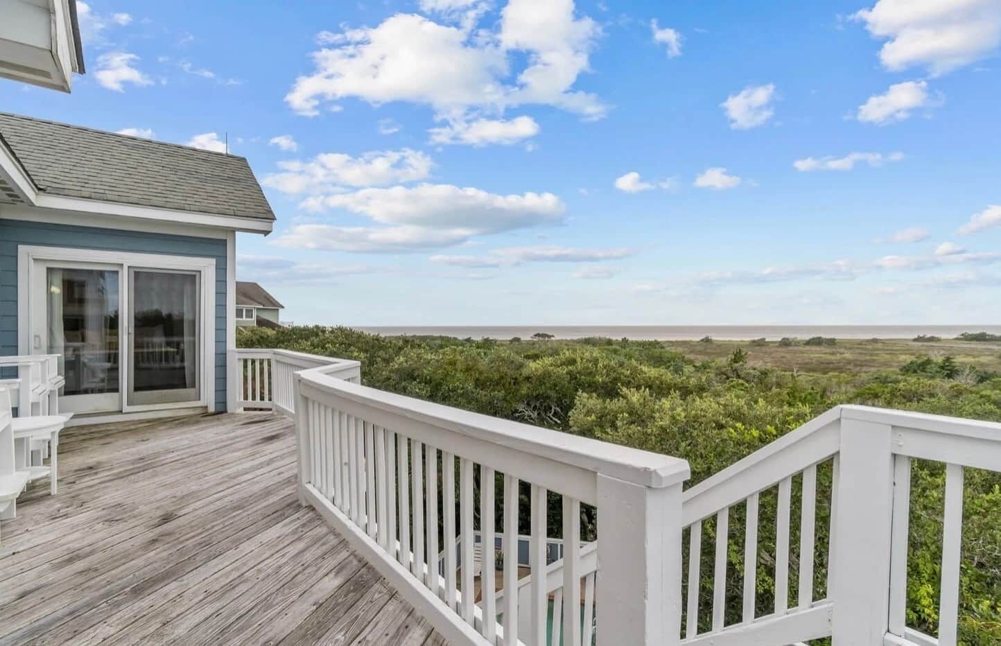 A wooden deck with a white railing, a glass door entrance, and a view of the beach and ocean.