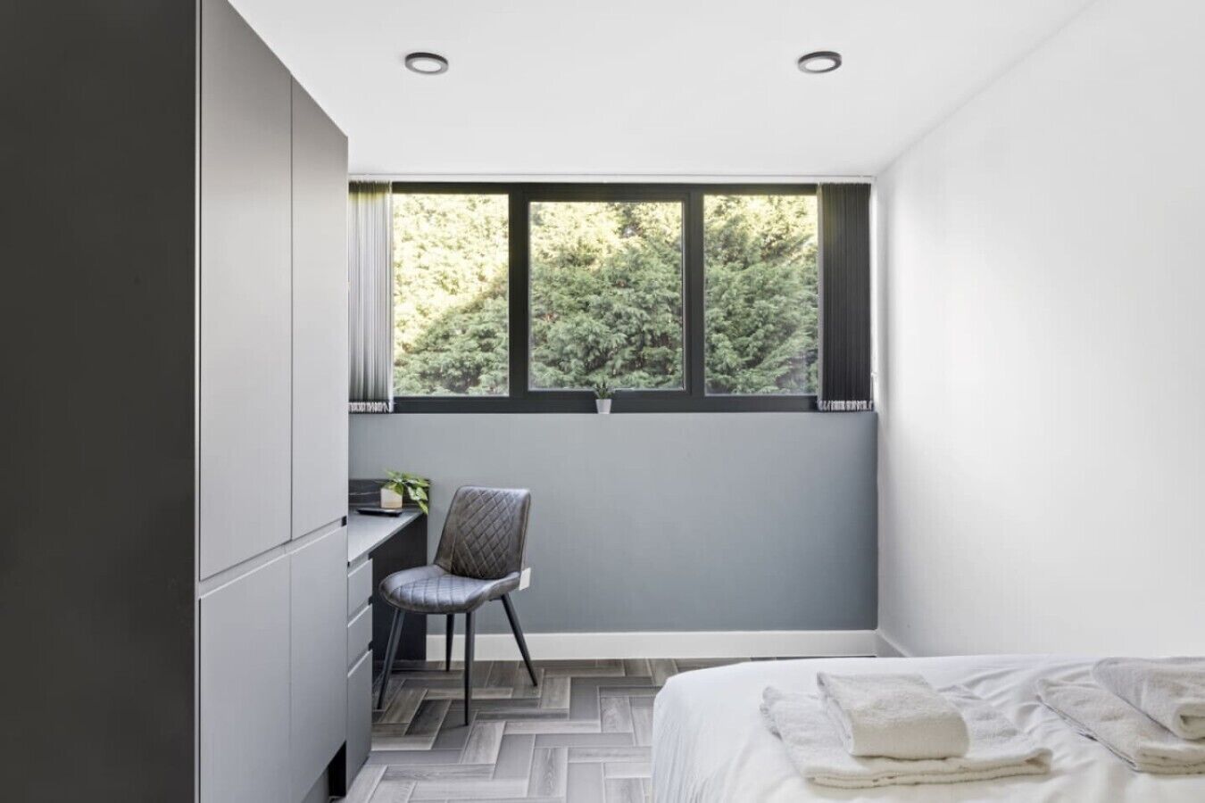 Une chambre à coucher avec un lit, un bureau et une chaise. Il y a une fenêtre donnant sur des arbres.