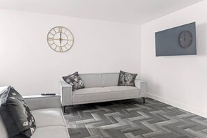 A modern living room with a grey sofa, two decorative pillows, a herringbone-patterned floor, a round wall clock, and a framed picture. - Brand New Spacious Studio in Beeston (Beeston)
