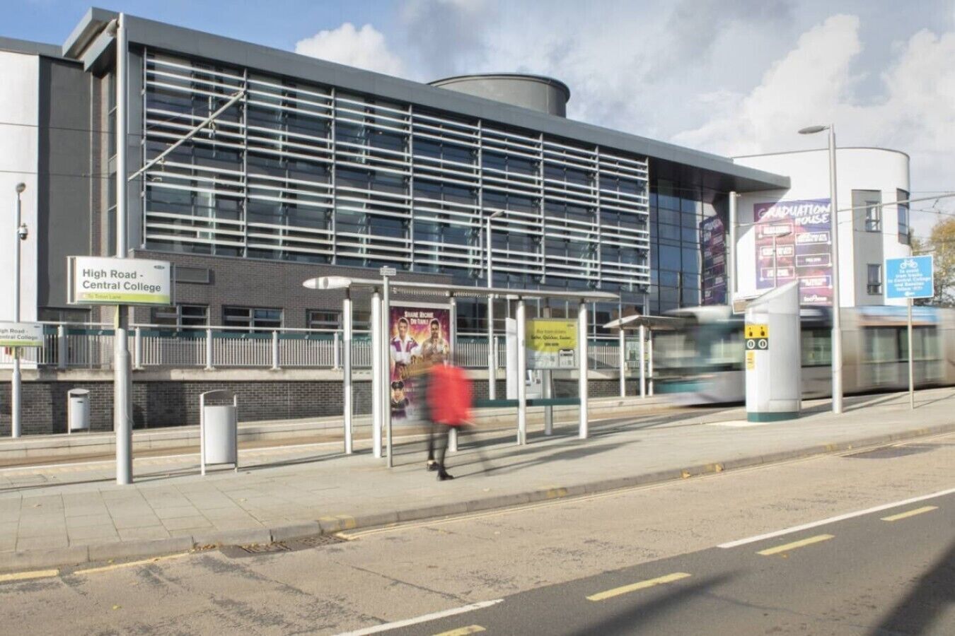 A modern building with a glass facade, a bus stop, and a sign for High Road - Central College.
