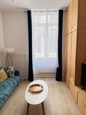 Una habitación con una cama, una mesita, un radiador, una ventana con cortinas y un armario de madera.