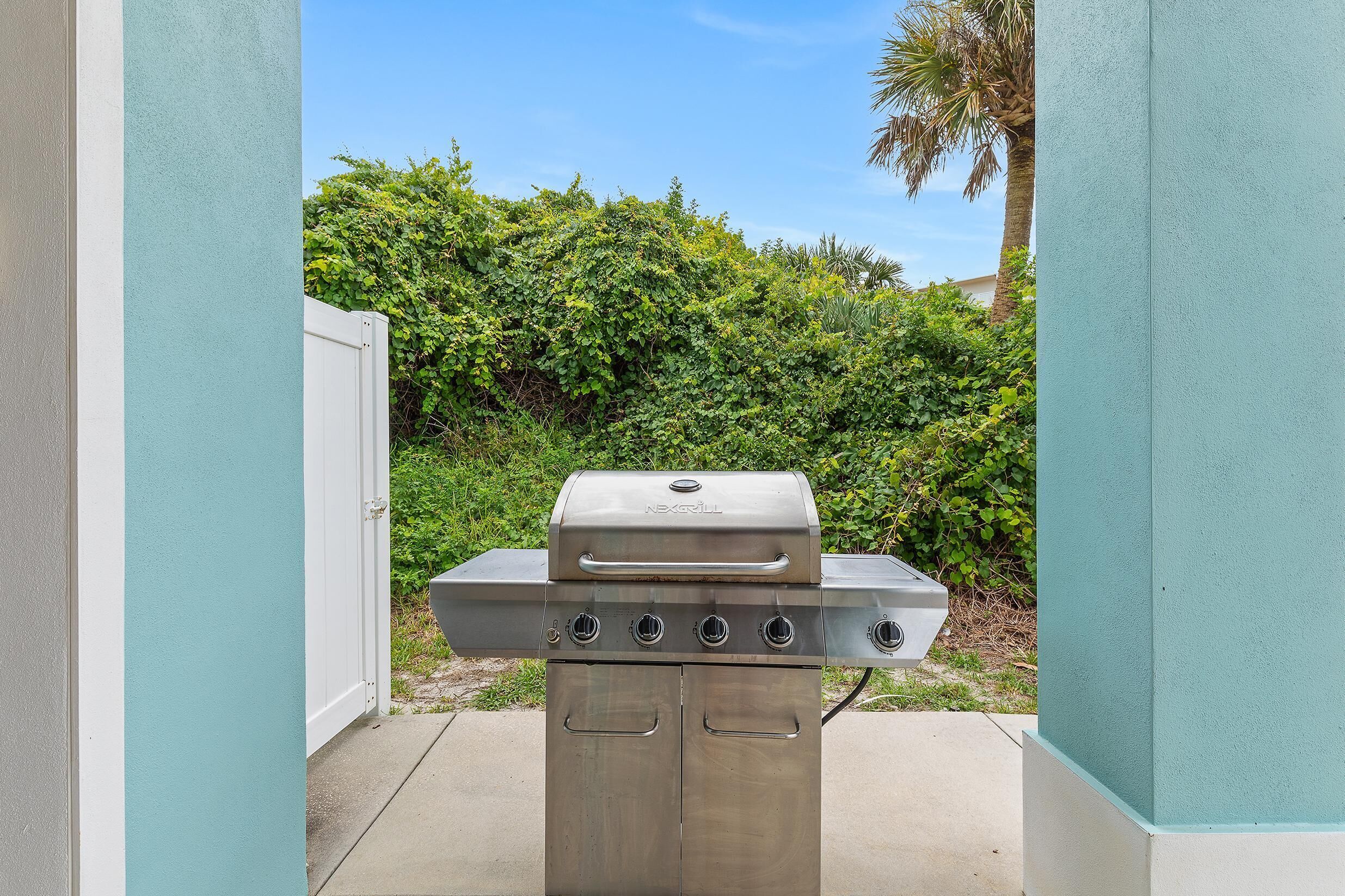 Una parrilla de acero inoxidable para exteriores con cuatro quemadores, ubicada en un acceso con una puerta blanca y un seto verde al fondo.