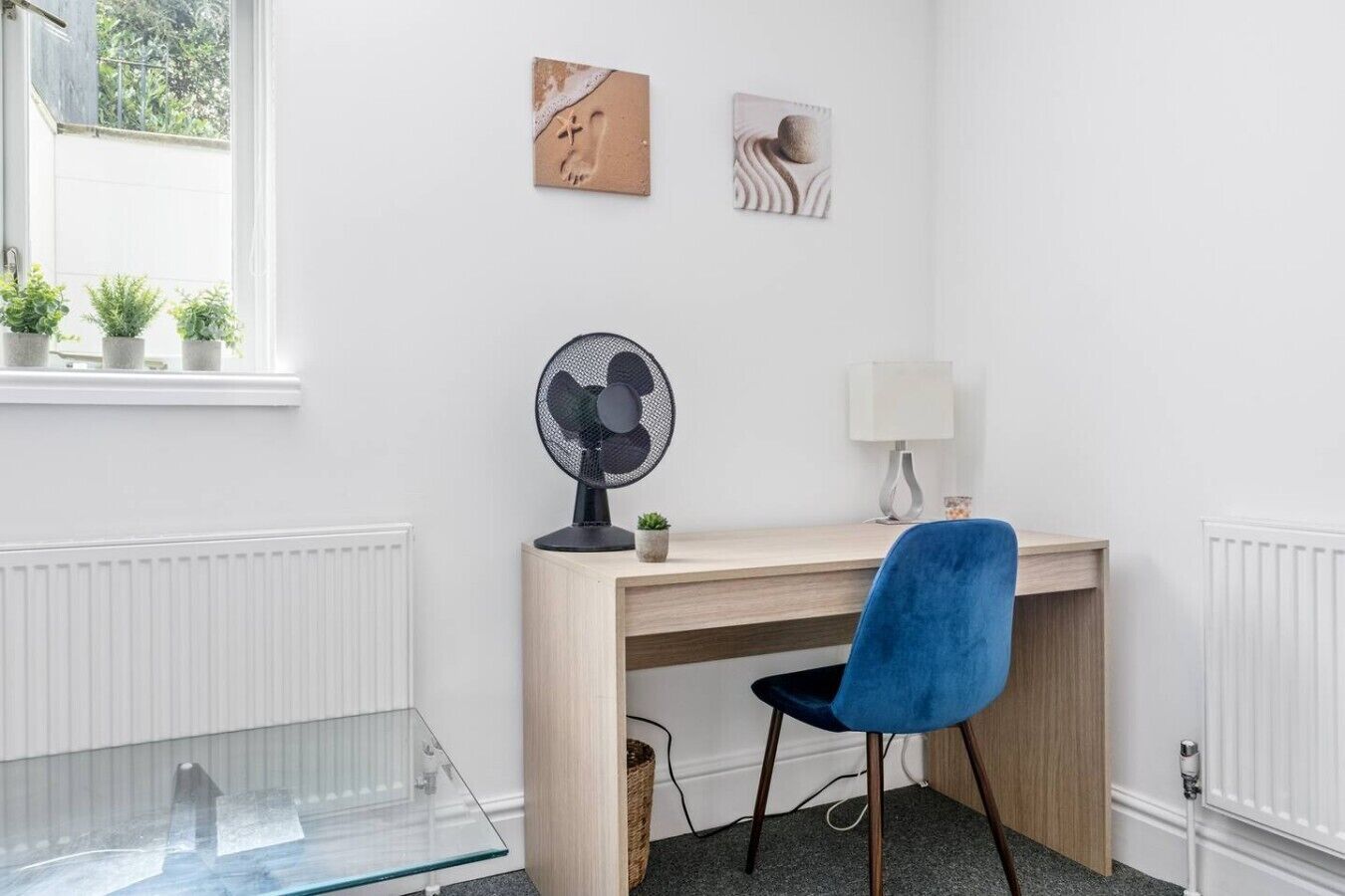 A room with a wooden desk, a blue chair, a standing fan, a lamp, and two wall-mounted pictures.