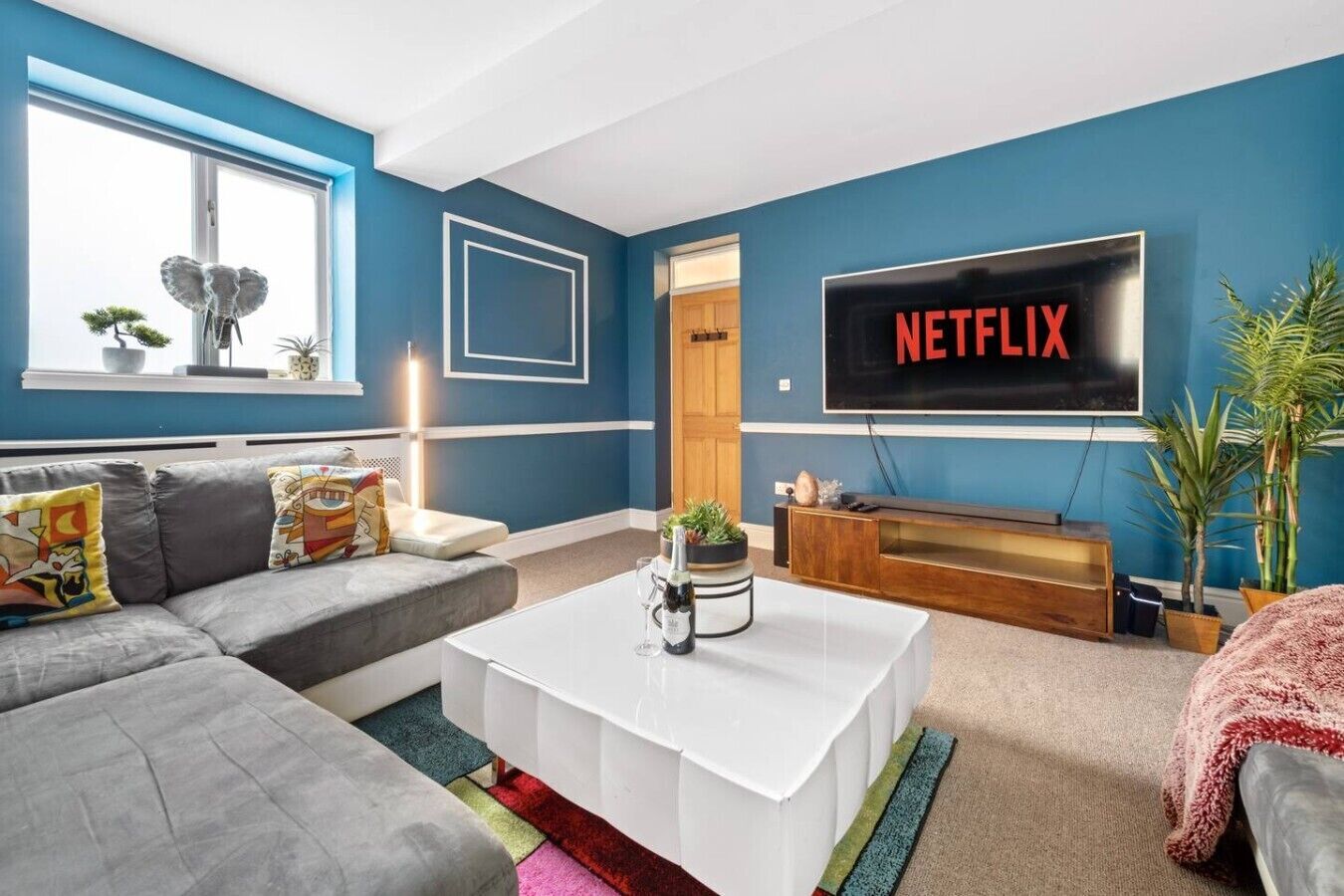Una sala de estar con un sofá gris, una mesa de centro blanca y un televisor que muestra 'NETFLIX' en letras rojas.