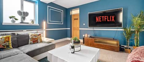 Una sala de estar con un sofá gris, una mesa de centro blanca y un televisor que muestra 'NETFLIX' en letras rojas.