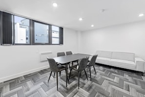 Ein modernes Zimmer mit Fischgrätparkett, einem Esstisch mit Stühlen, einem weißen Sofa und großen Fenstern mit Jalousien.