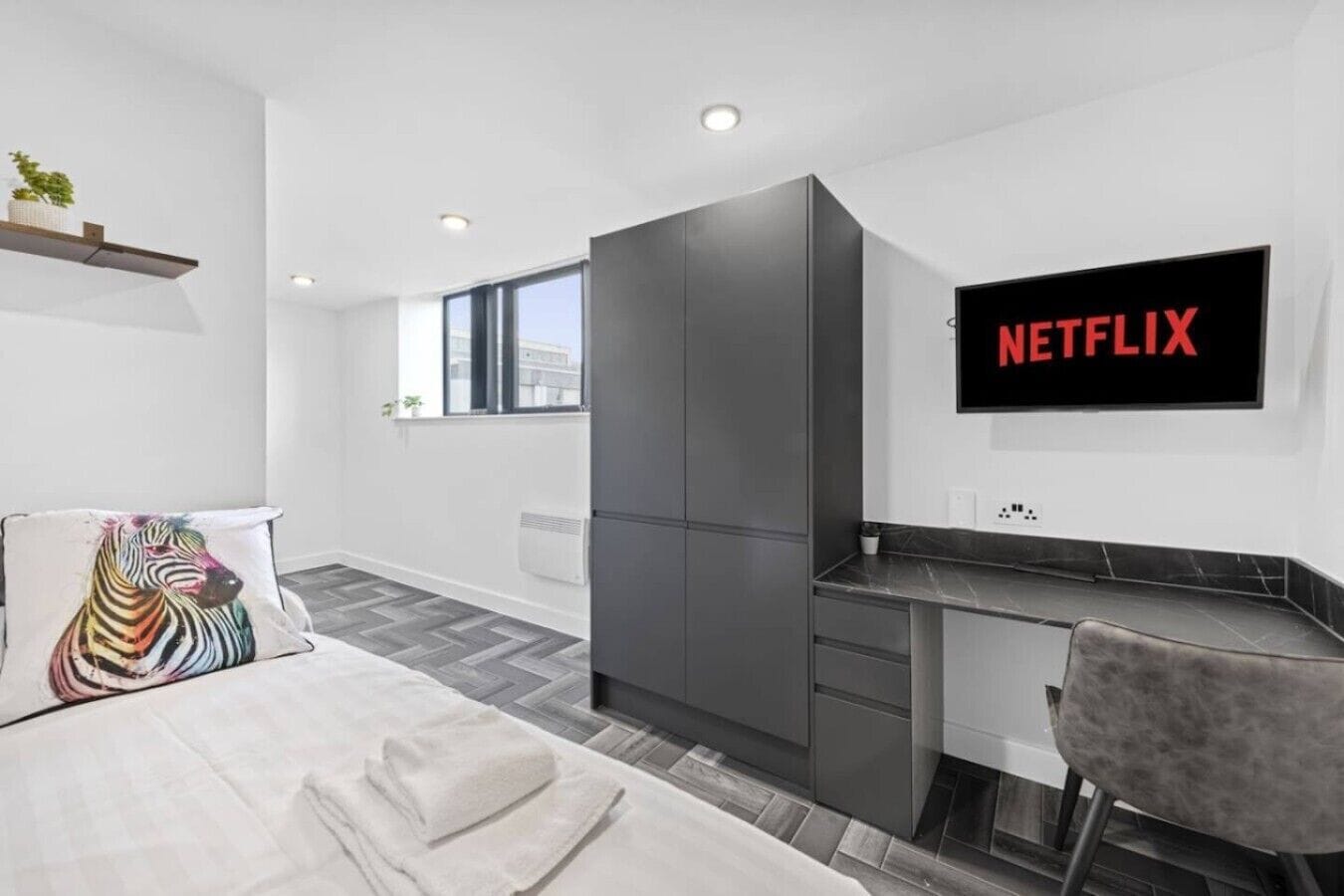 Ein modernes Hotelzimmer mit einem Bett, einem Schreibtisch, einem Stuhl, einem Kleiderschrank und einem an der Wand befestigten Fernseher mit dem Netflix-Logo.