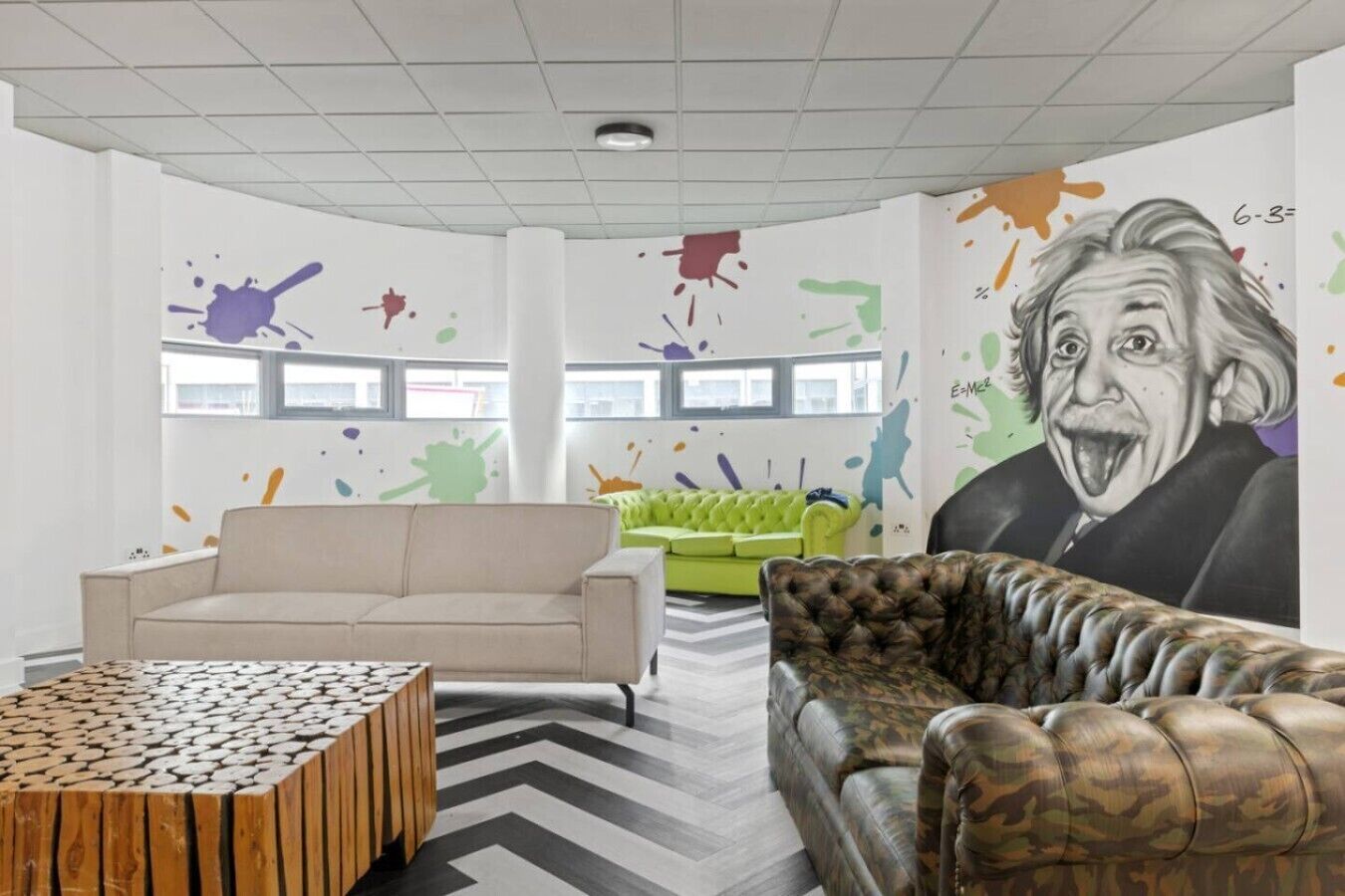Una sala moderna con un sofá beige, un sofá verde, un reposapiés estampado y un mural grande en la pared.