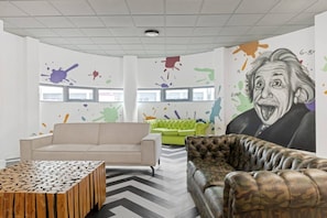 Ein modernes Wohnzimmer mit einem beigefarbenen Sofa, einem grünen Sofa, einem gemusterten Pouf und einem großen Wandgemälde.