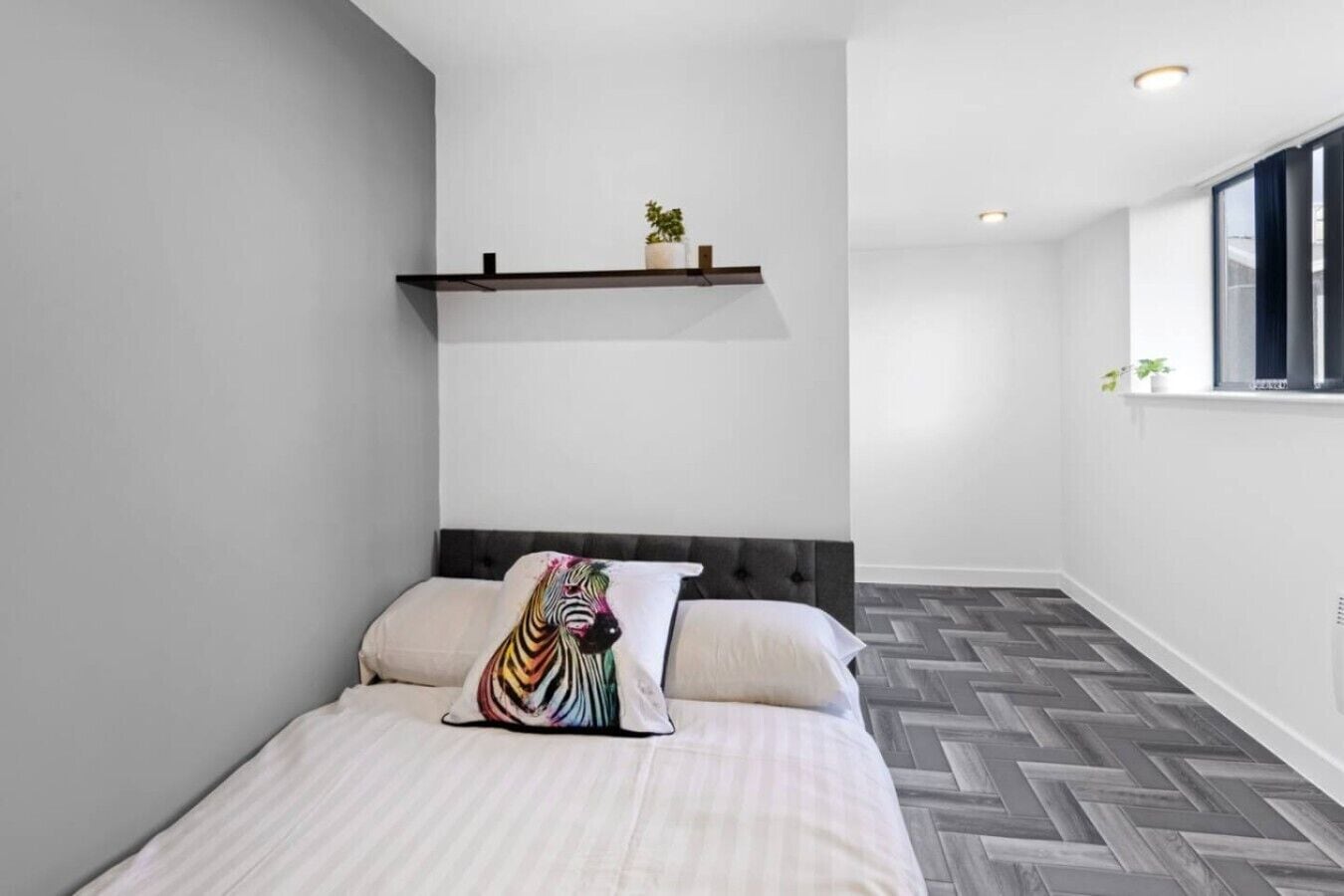 A modern bedroom with a bed, a zebra-patterned pillow, a wooden shelf with plants, and a herringbone-patterned floor.