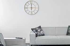 Un saló amb un sofà modern, un rellotge penjat a la paret i un coixí decoratiu.