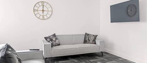 Una sala moderna con un sofá gris, dos almohadones decorativos, piso con patrón de espiga, un reloj de pared redondo y un cuadro enmarcado.